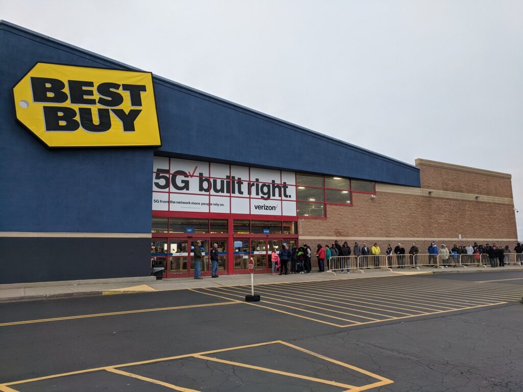 Façade bleue d'un magasin Best Buy au logo jaune, avec une longue file de clients derrière des barrières, tôt le matin, sur un parking presque vide