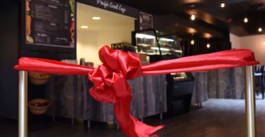 Ruban de satin rouge noué entre deux poteaux dorés devant le comptoir et la vitrine d’un café, juste avant son inauguration