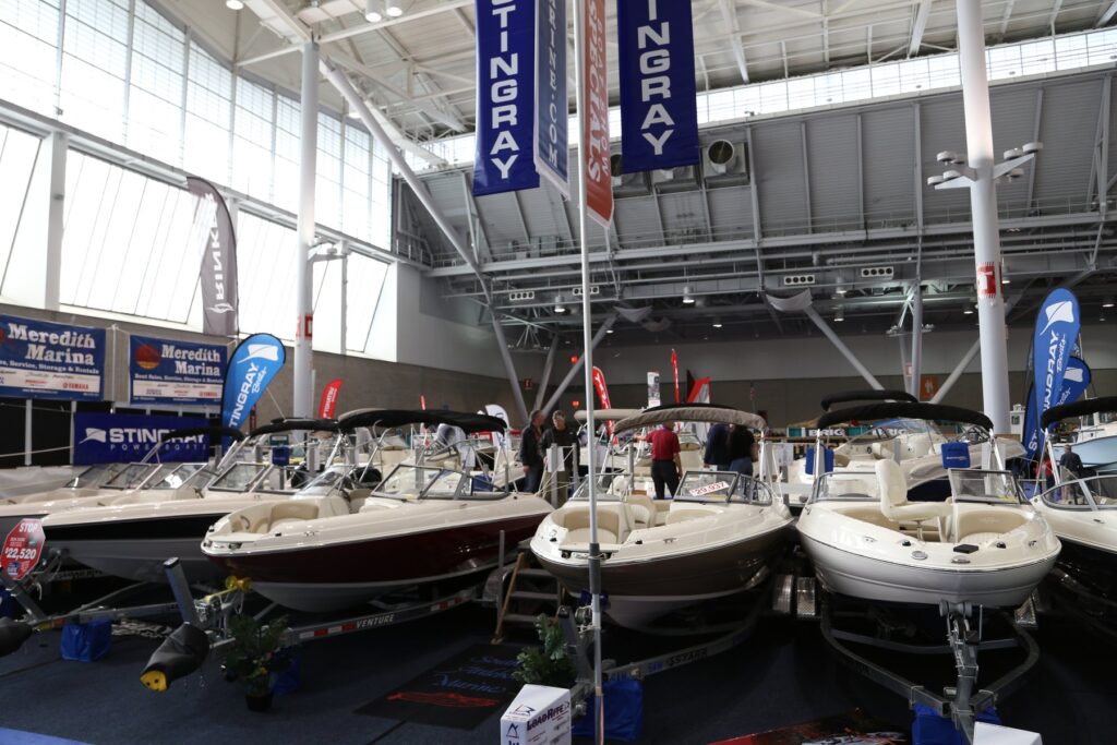 Rangée de bateaux à moteur de couleur crème sur leurs remorques, sous des oriflammes bleues Stingray, dans un hall vitré du New England Boat Show