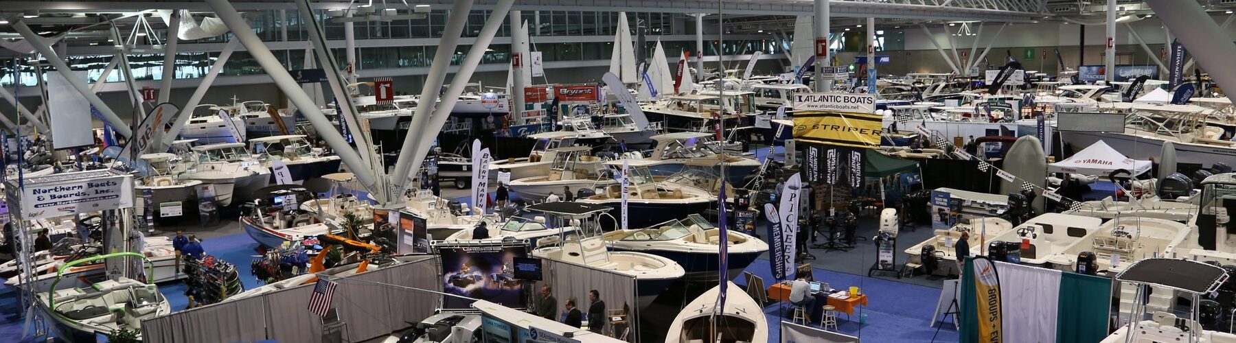 Vue en hauteur sur un hall d'exposition du New England Boat Show, avec des dizaines de bateaux à moteur et de voiliers sous une charpente métallique blanche et des stands sur une moquette bleue