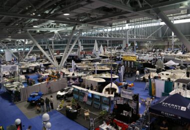 Vue en hauteur sur un hall d'exposition du New England Boat Show, avec des dizaines de bateaux à moteur et de voiliers sous une charpente métallique blanche et des stands sur une moquette bleue