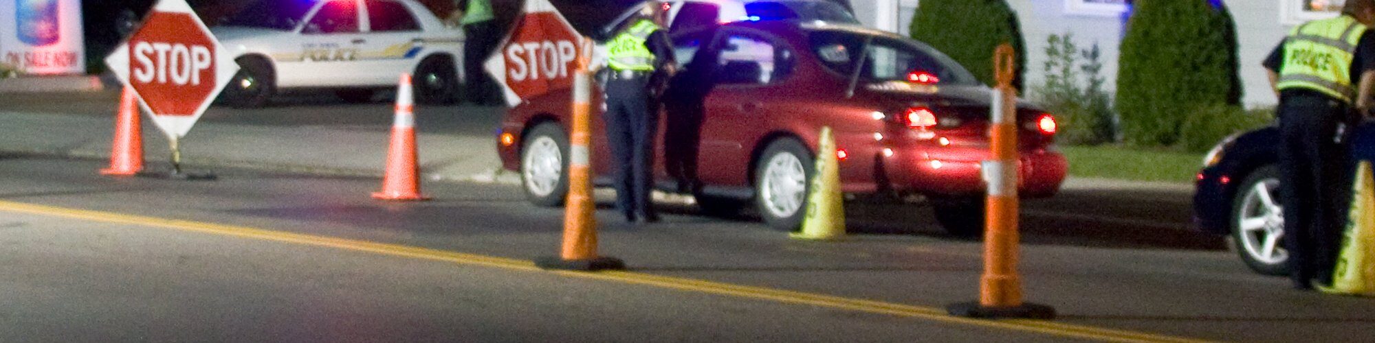 De nuit, des policiers en gilet jaune contrôlent des voitures entre des cônes orange et des panneaux Stop, devant des voitures de police aux gyrophares allumés