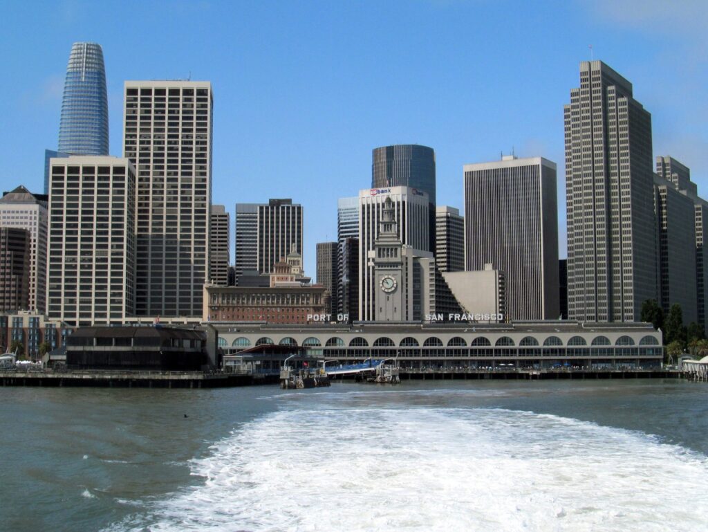 Le Ferry Building et ses quais vus depuis l'arrière d'un ferry qui s'éloigne, sillage blanc au premier plan, gratte-ciel du centre de San Francisco en arrière-plan