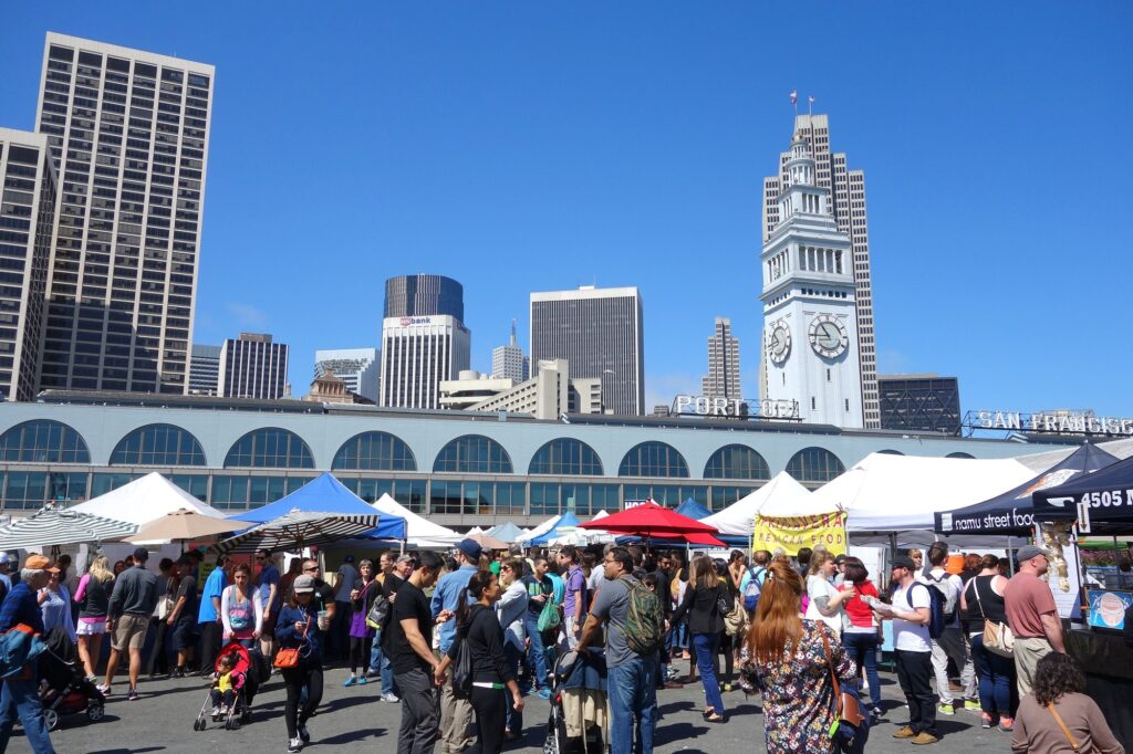 Foule entre les tentes du marché fermier du Ferry Building par grand soleil, tour de l'horloge et gratte-ciel du centre-ville en arrière-plan