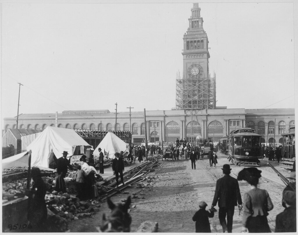 Photo en noir et blanc du Ferry Building en 1906, tour entourée d'échafaudages, tentes de sinistrés, gravats et tramway au premier plan