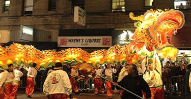 De nuit, la tête lumineuse orange et dorée d'un très long dragon et son corps illuminé ondulent au-dessus des porteurs en vestes blanches et pantalons rouges, devant une façade où se lit l'enseigne « Wayne's Liquors »