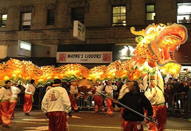 De nuit, la tête lumineuse orange et dorée d'un très long dragon et son corps illuminé ondulent au-dessus des porteurs en vestes blanches et pantalons rouges, devant une façade où se lit l'enseigne « Wayne's Liquors »