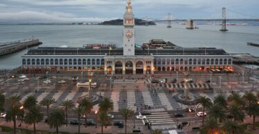 Vue en hauteur du Ferry Building de San Francisco au crépuscule, tour de l'horloge illuminée, Embarcadero bordé de palmiers, baie et Bay Bridge en arrière-plan