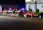 De nuit, des policiers en gilet jaune contrôlent des voitures entre des cônes orange et des panneaux Stop, devant des voitures de police aux gyrophares allumés