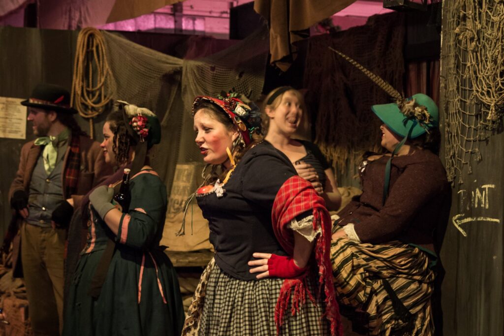 Comédiennes et comédiens en costumes victoriens, châle à carreaux et chapeaux, dans un décor de rue de Londres à la Dickens Fair