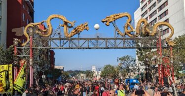 Foule massée sous le portique de Chinatown, une arche métallique surmontée de deux grands dragons dorés, avec des bannières jaunes de danse du lion au premier plan