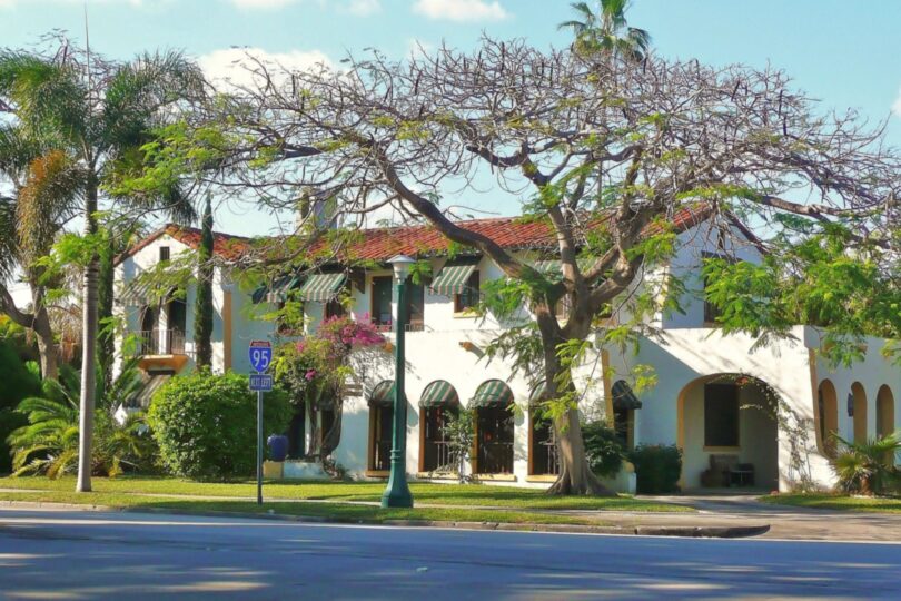 Maison blanche de style méditerranéen à toit de tuiles rouges, stores rayés verts et arcades, sous un grand arbre et des palmiers, dans une rue de Miami Shores