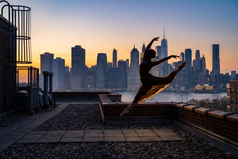 Silhouette à contre-jour d'une danseuse en plein saut sur le toit d'un immeuble, devant les gratte-ciel de Manhattan au crépuscule
