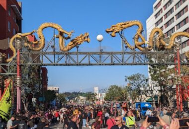 Foule massée sous le portique de Chinatown, une arche métallique surmontée de deux grands dragons dorés, avec des bannières jaunes de danse du lion au premier plan