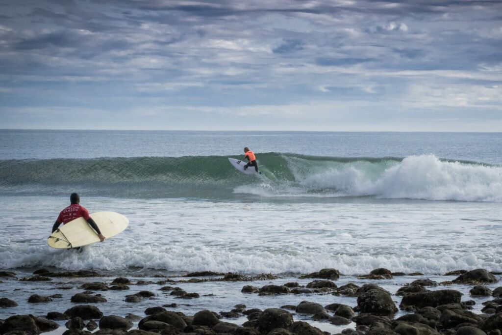Surfeur en lycra orange sur une vague qui déroule vers la droite devant une plage de galets à Rincon, pendant qu'un concurrent du Rincon Classic entre dans l'eau, sa planche sous le bras