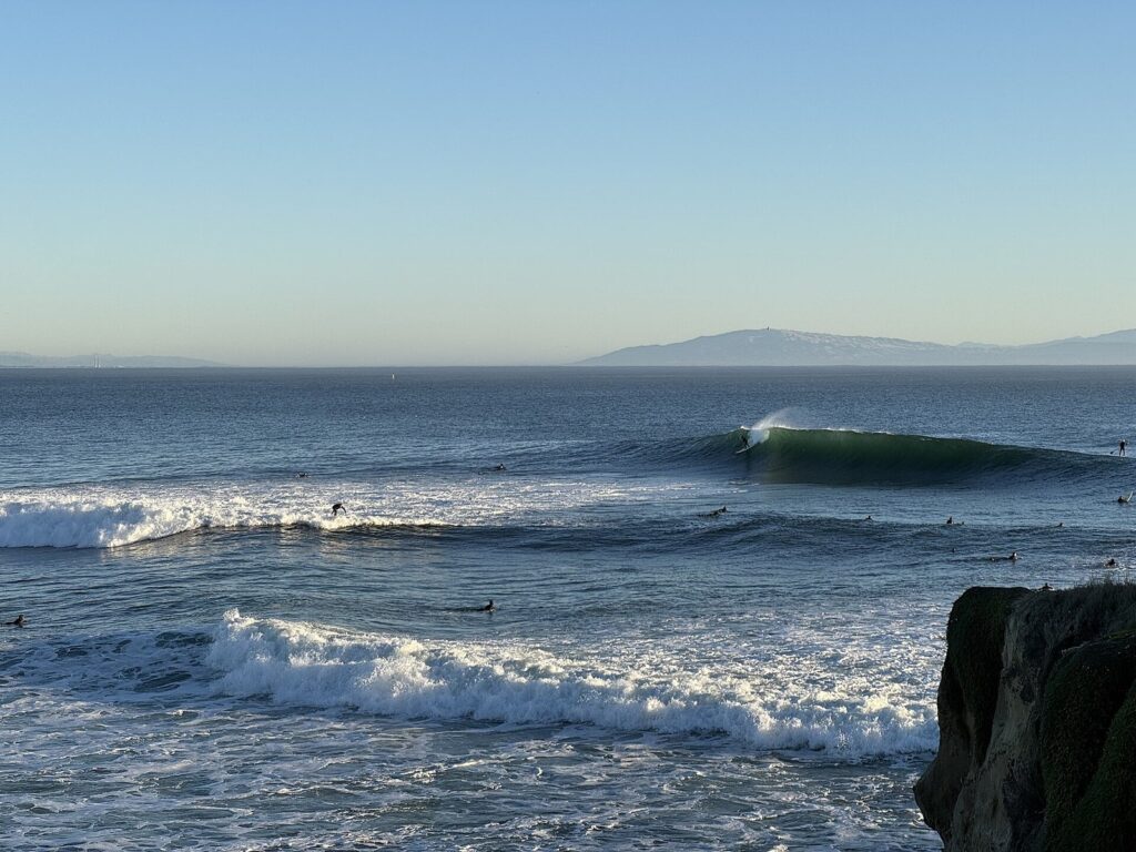Surfeur sur une vague verte à Steamer Lane, à Santa Cruz, avec une vingtaine de surfeurs au large, une falaise au premier plan et la baie de Monterey en arrière-plan