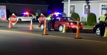 De nuit, des policiers en gilet jaune contrôlent des voitures entre des cônes orange et des panneaux Stop, devant des voitures de police aux gyrophares allumés