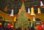 Grand sapin illuminé sous la verrière du jardin d'hiver du Bellagio, entouré de cloches géantes dorées et argentées suspendues, avec des visiteurs
