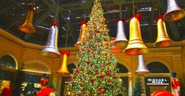 Grand sapin illuminé sous la verrière du jardin d'hiver du Bellagio, entouré de cloches géantes dorées et argentées suspendues, avec des visiteurs