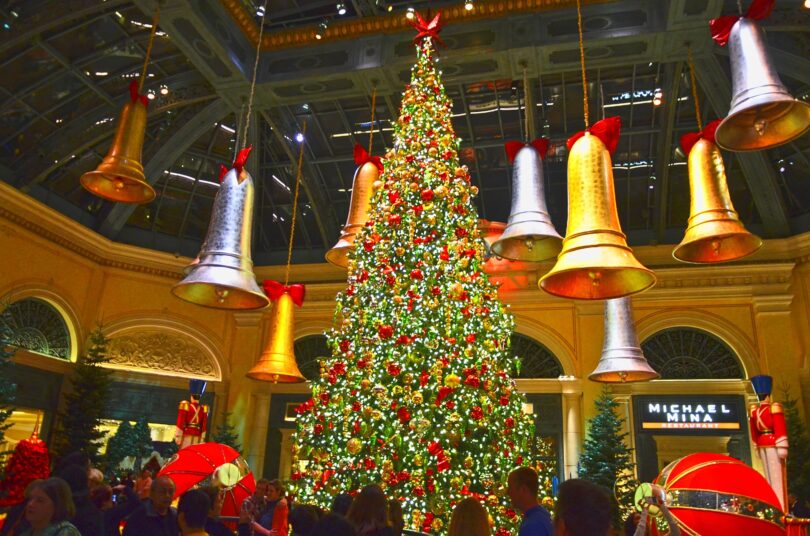 Grand sapin illuminé sous la verrière du jardin d'hiver du Bellagio, entouré de cloches géantes dorées et argentées suspendues, avec des visiteurs
