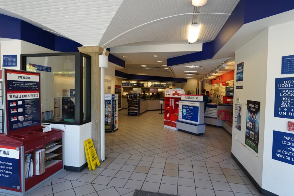 Entrée d'un bureau de poste américain aux couleurs bleu, blanc et rouge de l'USPS, avec un présentoir d'emballages Priority Mail à gauche, des rayons de fournitures d'expédition et les guichets au fond