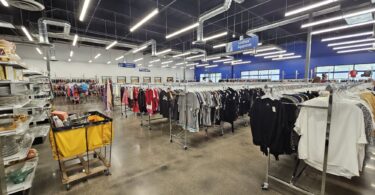 Grand magasin Goodwill aux plafonds noirs et néons : longues rangées de portants de vêtements, panneau bleu « Ladies’ Apparel », étagères de vaisselle et de verres à gauche, un chariot de tri jaune au premier plan