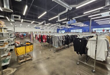 Grand magasin Goodwill aux plafonds noirs et néons : longues rangées de portants de vêtements, panneau bleu « Ladies’ Apparel », étagères de vaisselle et de verres à gauche, un chariot de tri jaune au premier plan