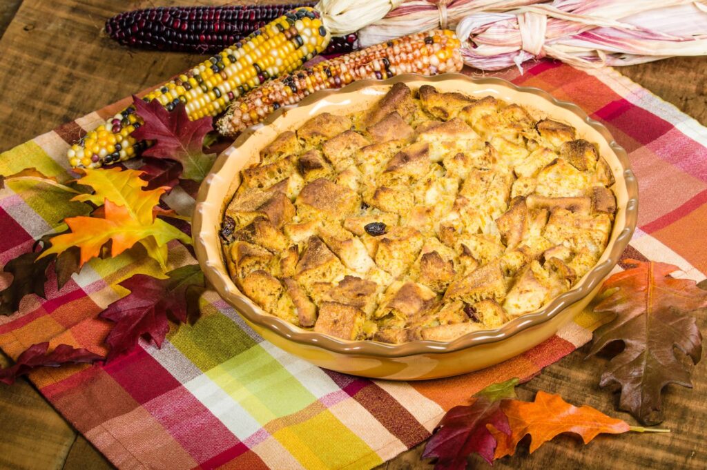 Bread pudding aux raisins secs dans un plat à tarte en céramique, posé sur une nappe à carreaux avec des épis de maïs décoratifs et des feuilles d'automne