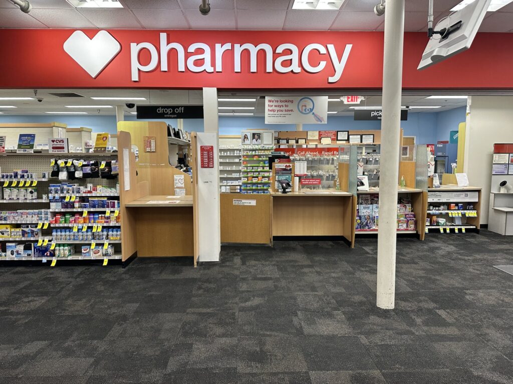 Comptoir de pharmacie d'un CVS sous une grande enseigne rouge Pharmacy, avec les guichets de dépôt et de retrait des ordonnances, entre des rayons de produits en vente libre