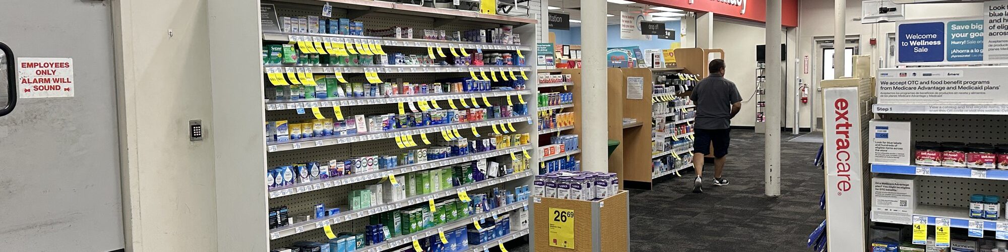 Allée d'un CVS bordée de rayons de médicaments en vente libre, avec l'enseigne rouge de la pharmacie au fond