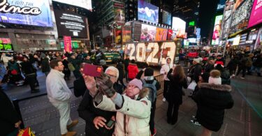 Foule de nuit à Times Square, entre les écrans géants, devant les grands chiffres lumineux « 2023 » installés pour le réveillon ; un couple se prend en photo