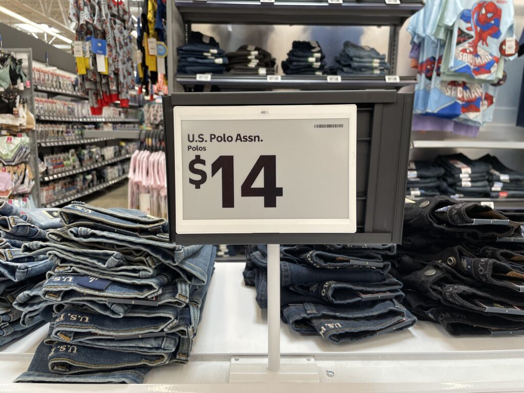 Étiquette de prix à écran électronique gris affichant « U.S. Polo Assn. Polos, $14 », posée au-dessus de piles de jeans dans le rayon vêtements d'un magasin Walmart
