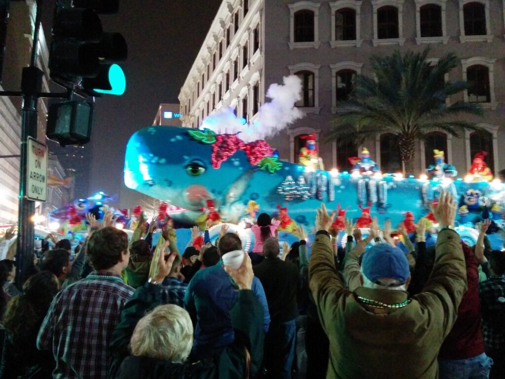 De nuit, une immense baleine bleue illuminée, crachant un jet de fumée blanche et chargée de cavaliers costumés, avance devant un immeuble ancien ; au premier plan, la foule lève les bras