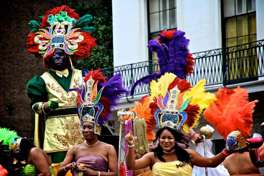 Sur un char, des femmes en robes de satin et immenses coiffes de plumes rouges, violettes, jaunes et orange, derrière elles un homme en costume vert et or ; l'une tient une noix de coco pailletée violet, vert et or