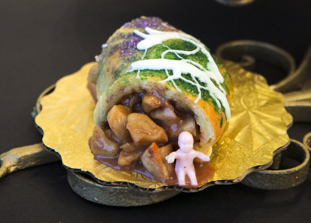 Part de king cake roulée, glacée de blanc et saupoudrée de sucre violet, vert et or, à la garniture en gros morceaux, avec une petite figurine de bébé en plastique rose posée devant, sur un carton doré