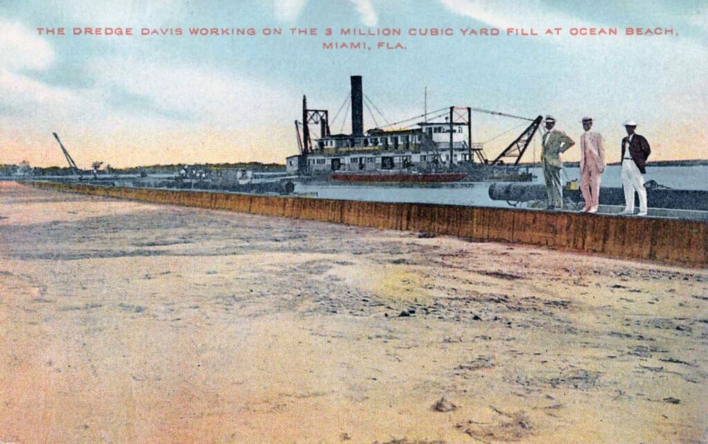 Carte postale colorisée vers 1915 : la drague à vapeur Davis sur la baie, derrière un long remblai de sable fraîchement déversé où posent trois hommes en costume clair