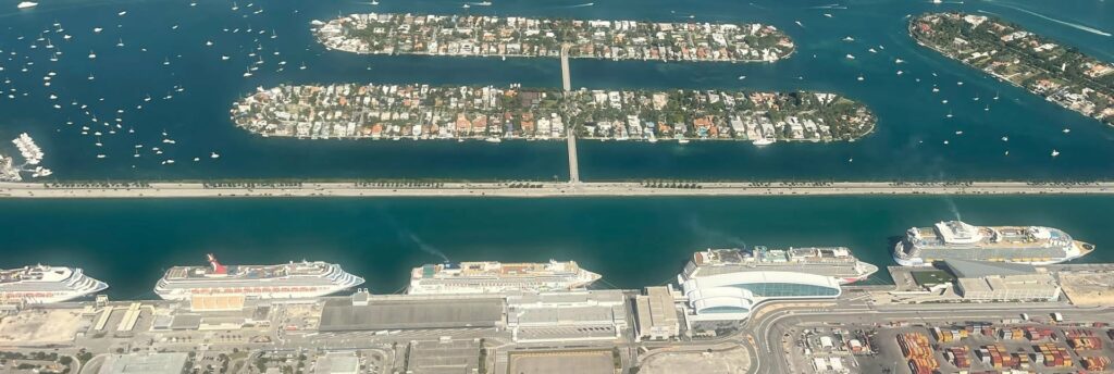 Vue aérienne de PortMiami : cinq paquebots de croisière alignés à quai devant les terminaux de Dodge Island, avec au-delà de la chaussée deux îles résidentielles artificielles dans la baie de Biscayne
