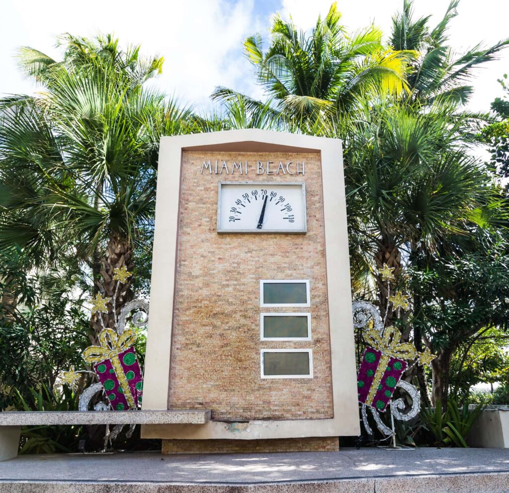Grand thermomètre à cadran marqué Miami Beach, gradué de 20 à 110 degrés Fahrenheit, sur une stèle en pierre beige devant des palmiers