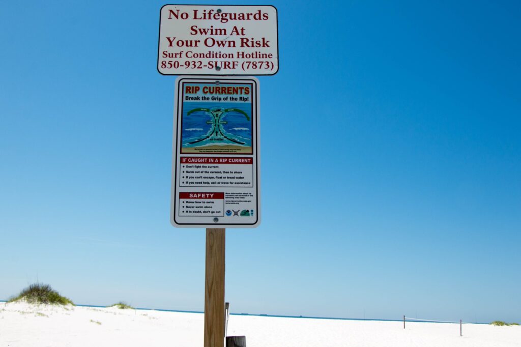 Sur une plage de sable blanc, deux panneaux sur un poteau : No Lifeguards, Swim At Your Own Risk, avec le numéro d'information sur l'état de la mer, et Rip Currents, Break the Grip of the Rip!, qui montre comment sortir d'un courant