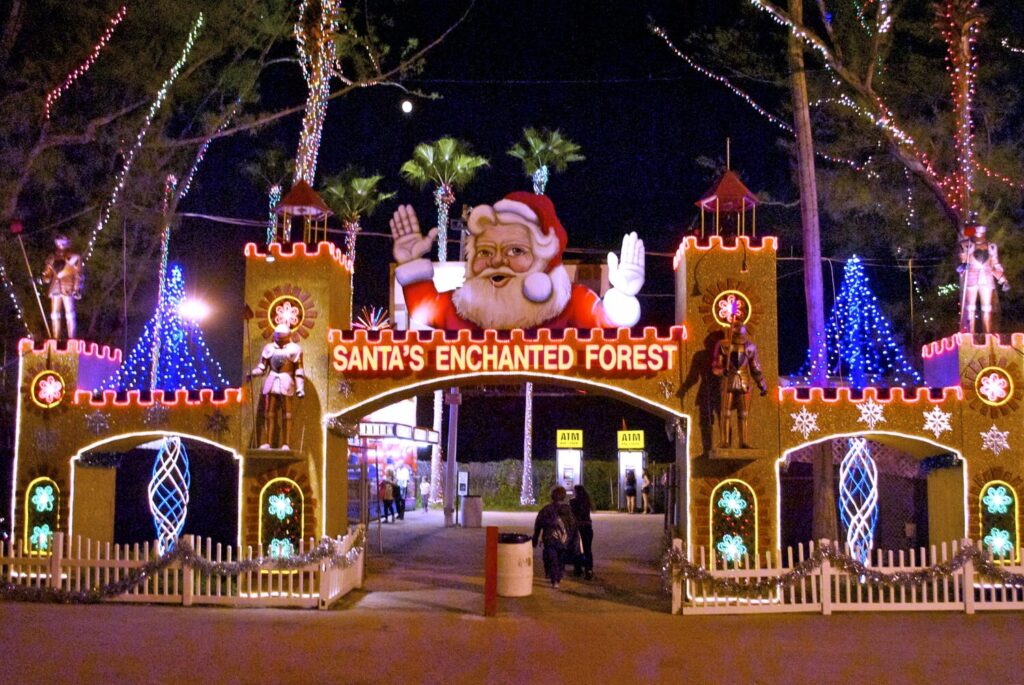 Portail d'entrée de fête foraine en forme de château illuminé, surmonté d'un grand Père Noël qui salue, arbres et palmiers couverts de guirlandes lumineuses la nuit