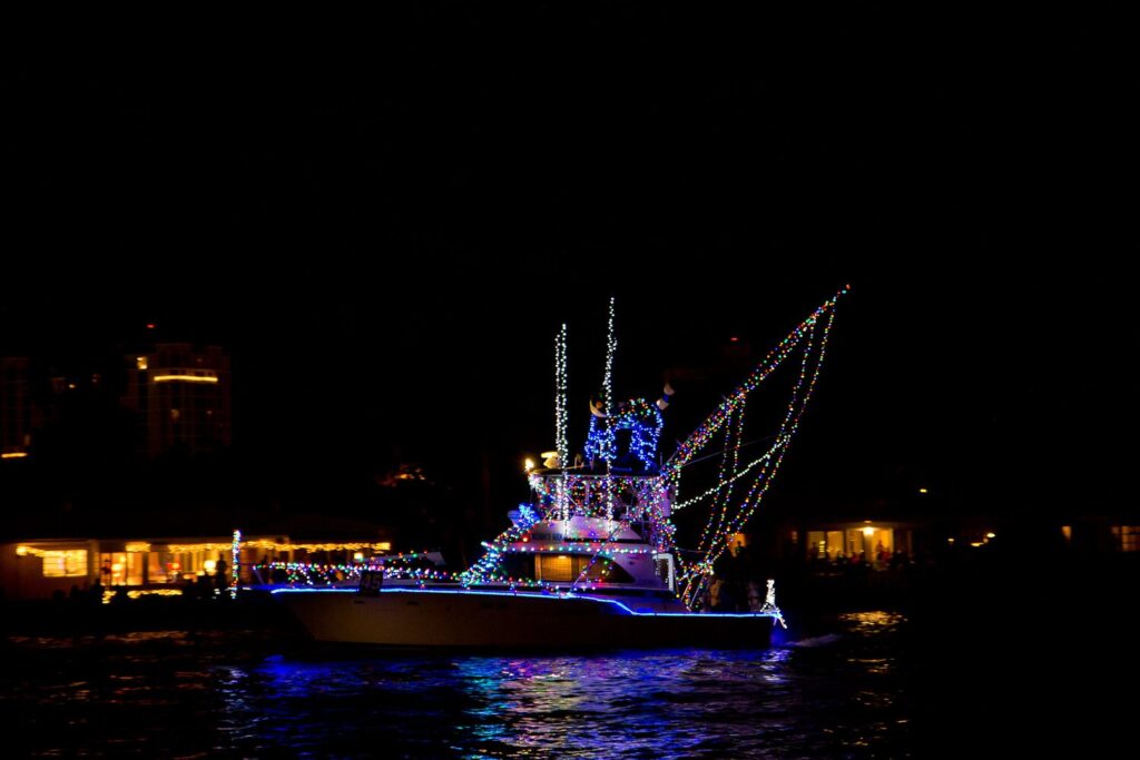 Bateau de pêche sportive couvert de guirlandes lumineuses bleues et blanches naviguant de nuit, reflets colorés sur l'eau et immeubles éclairés au loin
