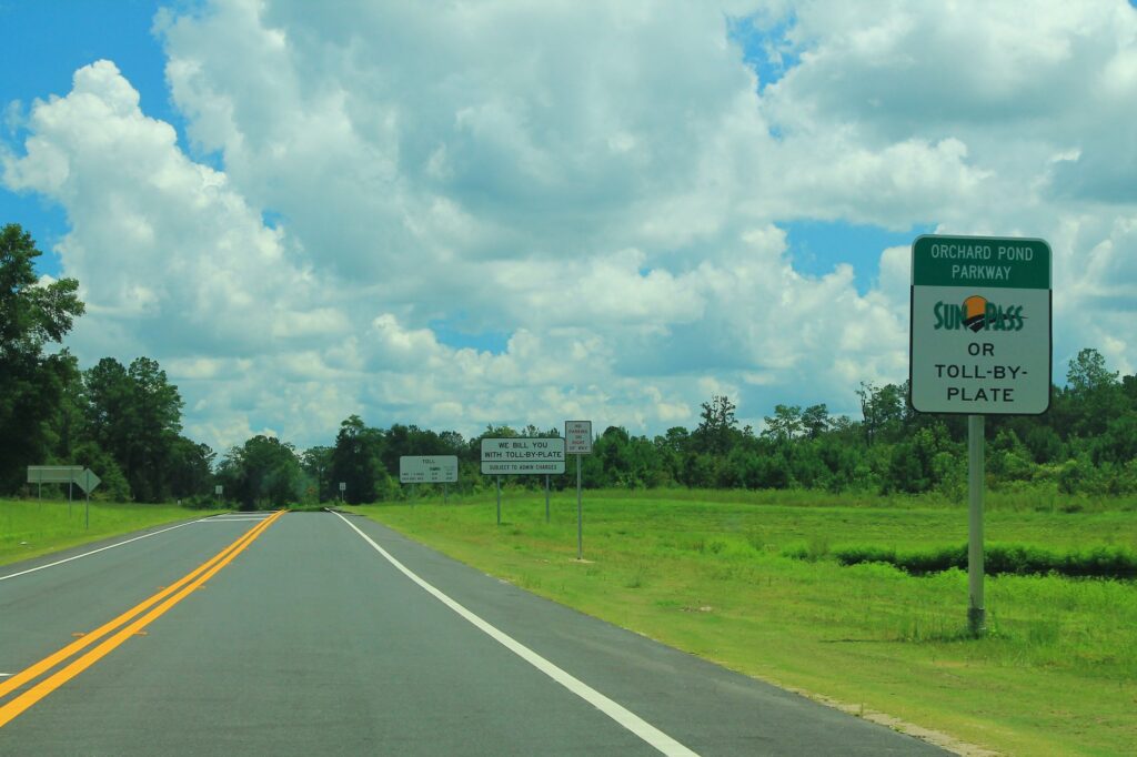 Route à deux voies entre des pelouses, avec à droite un panneau vert Orchard Pond Parkway portant le logo SunPass et la mention Or Toll-By-Plate