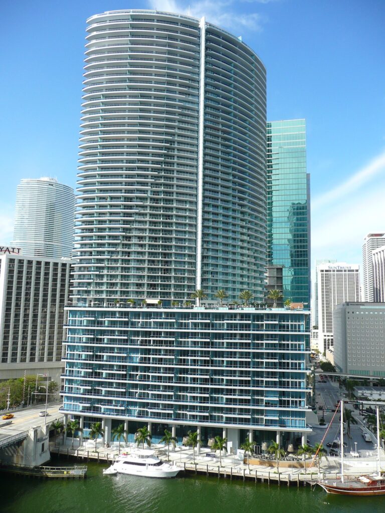 Gratte-ciel courbe aux balcons blancs, posé sur un socle vitré bleu-vert, au bord de la Miami River à Downtown Miami, avec une rangée de palmiers et un yacht à quai à son pied