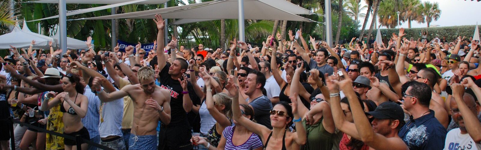 Une foule en maillot de bain et en tenue d'été lève les bras sous des voiles d'ombrage blanches et des palmiers, au bord d'une piscine, pendant une pool party de la Winter Music Conference à Miami