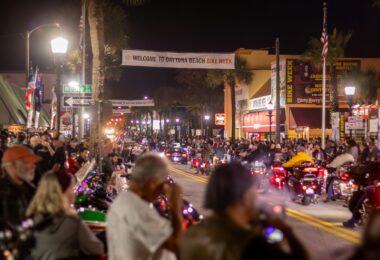 Main Street à Daytona Beach la nuit, sous une banderole Welcome to Daytona Beach Bike Week, avec des motos feux allumés au milieu d'une foule de motards et de badauds