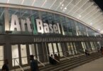 Façade vitrée du Miami Beach Convention Center éclairée de nuit, avec les grandes lettres Art Basel au-dessus des portes, des visiteurs assis sur les marches