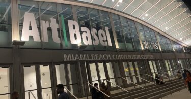 Façade vitrée du Miami Beach Convention Center éclairée de nuit, avec les grandes lettres Art Basel au-dessus des portes, des visiteurs assis sur les marches