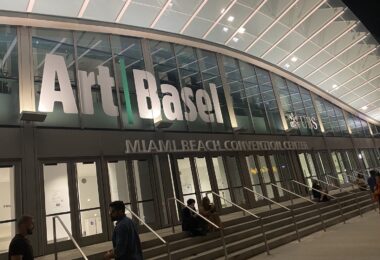Façade vitrée du Miami Beach Convention Center éclairée de nuit, avec les grandes lettres Art Basel au-dessus des portes, des visiteurs assis sur les marches