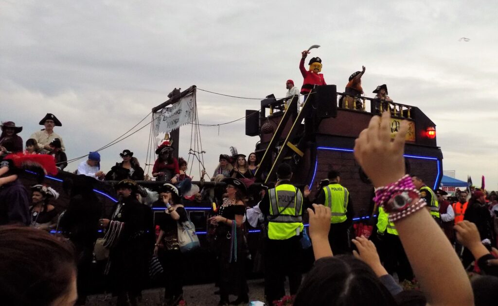 Un char en forme de navire pirate entouré de membres d'une krewe en costume de pirate, tricornes et colliers de perles, pendant que des spectateurs lèvent les bras pour attraper des colliers