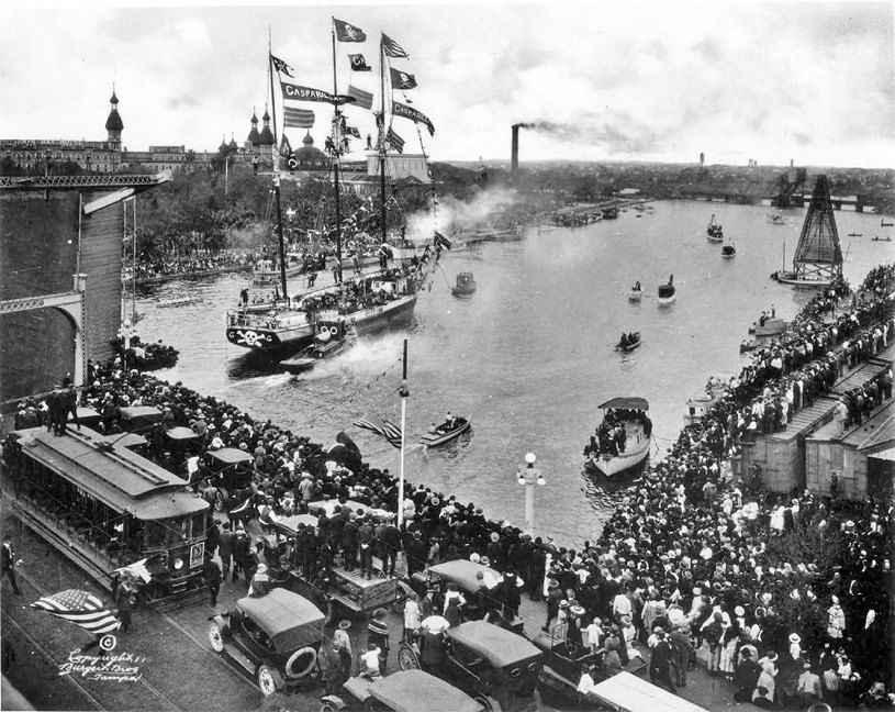 Photo ancienne en noir et blanc d'un trois-mâts pavoisé de drapeaux pirates remontant la Hillsborough River à Tampa, devant une foule massée sur les quais, avec un tramway et des voitures anciennes au premier plan et les minarets du Tampa Bay Hotel au loin